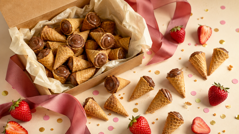 Open kraft gift box spilling chocolate-filled mini waffle cones with a berry-pink ribbon